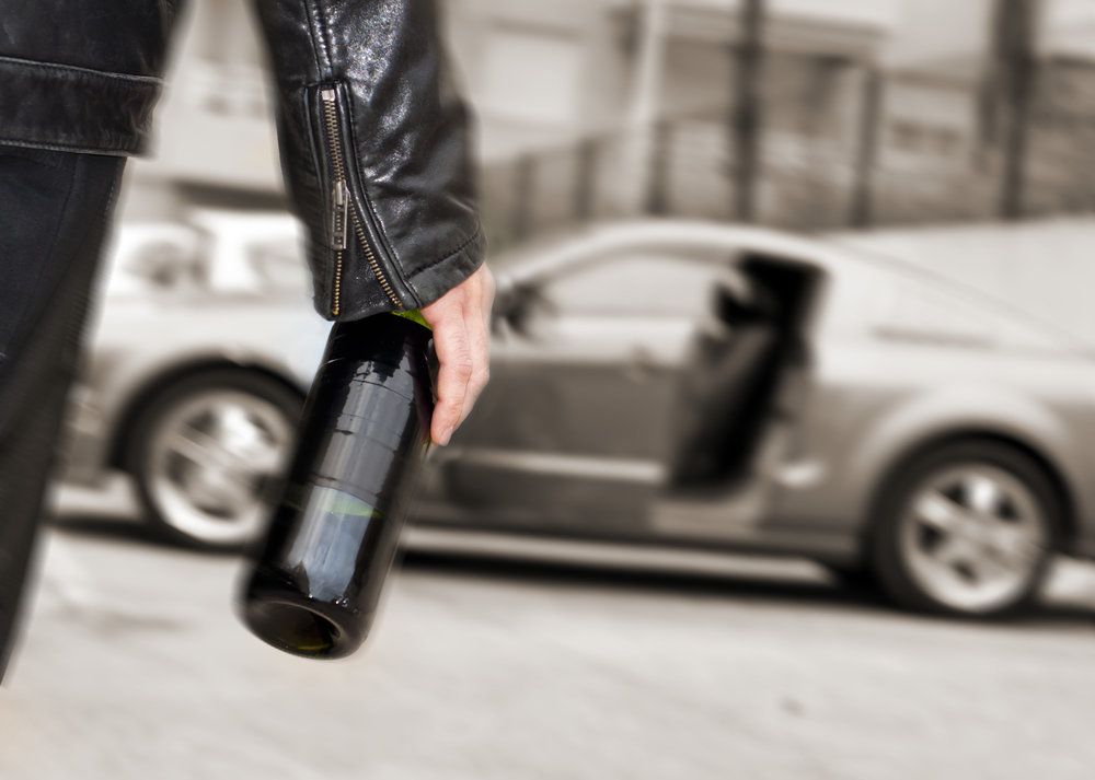 Man in leather coat walking towards car with wine bottle in his hand